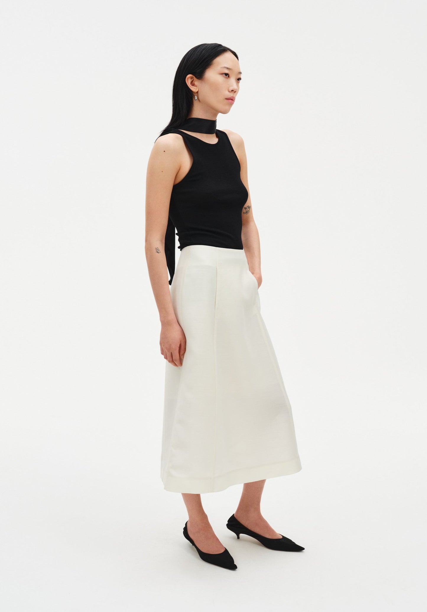 A woman stands in a three-quarter side pose against a plain white background, wearing a black sleeveless top with a high neckline and choker-style detail, paired with a cream-colored midi skirt and black pointed heels. Her dark shoulder-length hair is worn down, and she looks slightly to the side with a neutral expression.
