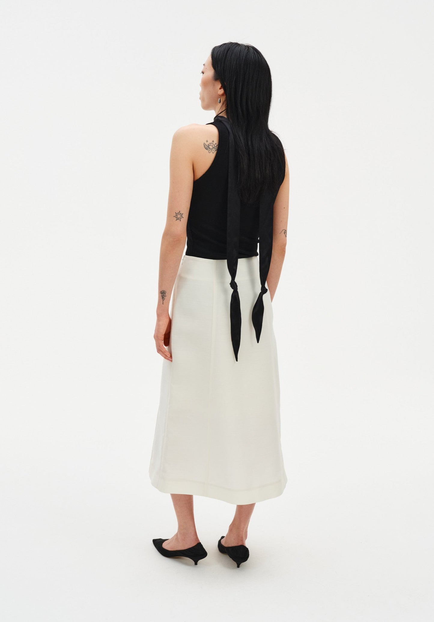 Rear three-quarter view of a woman against a plain white background, wearing a black sleeveless top with a long tie detail draping down the back, paired with a cream-colored midi skirt and black pointed heels. Her dark shoulder-length hair is worn down, and small tattoos are visible on her arms and upper back.
