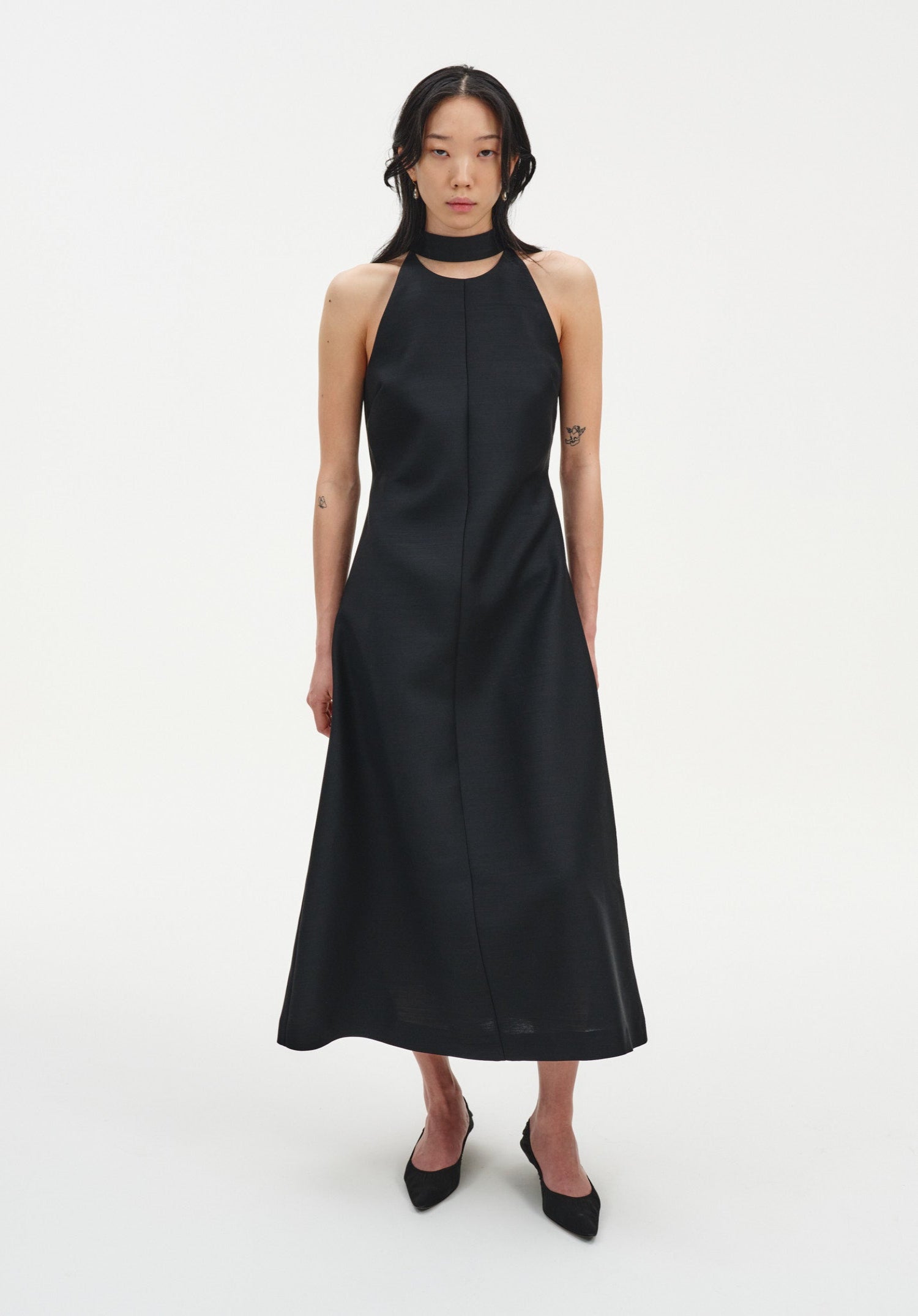 Full-length front view of a model wearing a sleeveless black midi dress with a choker-style neckline and A-line silhouette, paired with black pointed heels.
