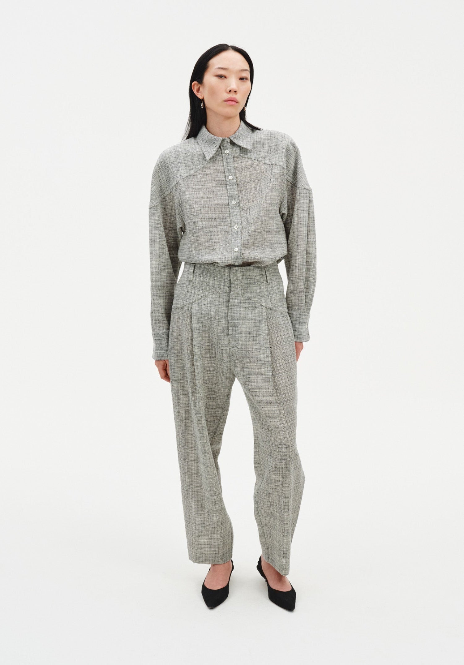 Full-length front view of a woman wearing a grey textured button-up shirt tucked into matching high-waisted trousers with soft pleats, styled with black pointed heels against a minimal white studio background.
