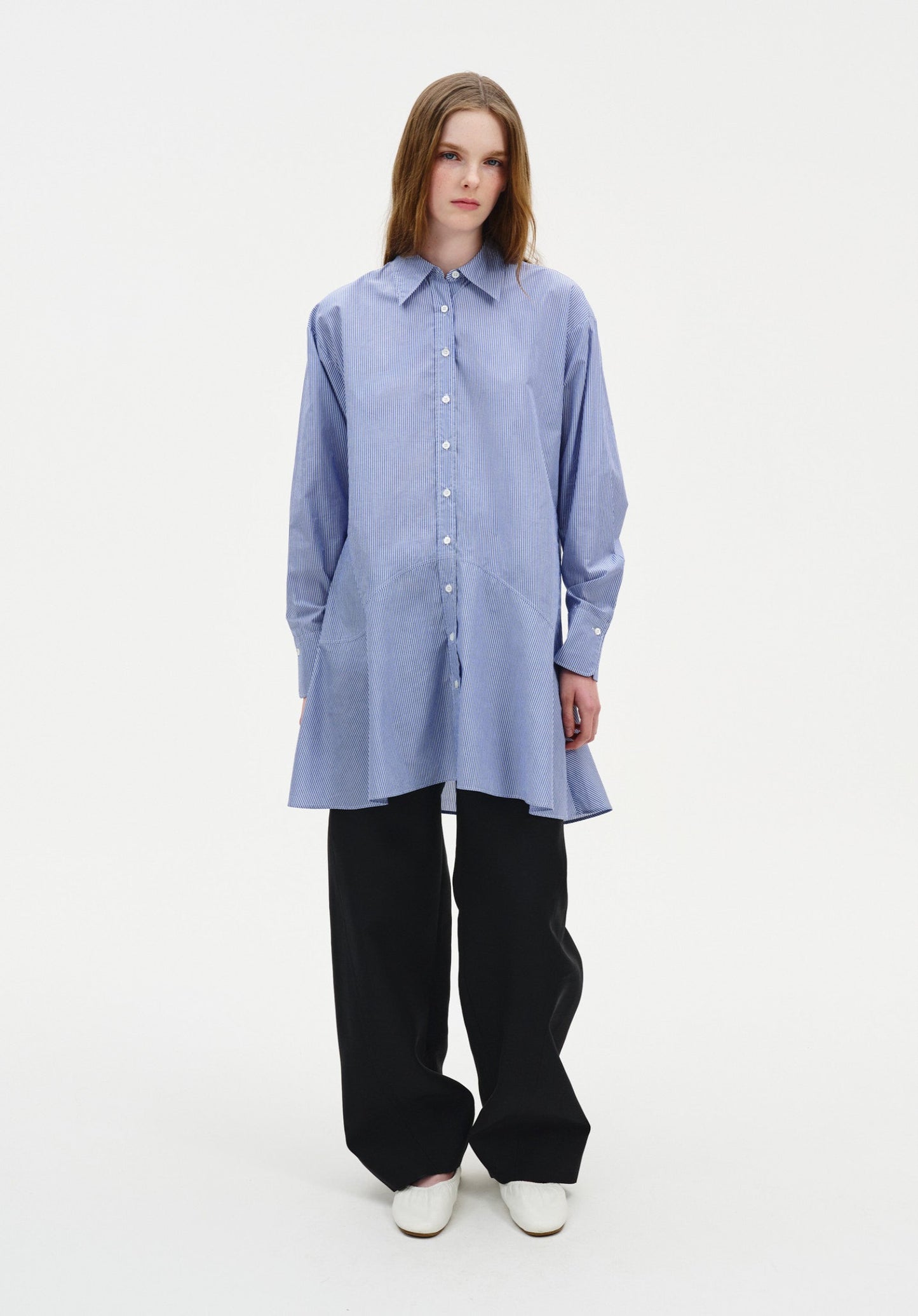 A woman stands facing forward against a plain white background, wearing a light blue striped button-up shirt with a flared hem and long sleeves, paired with loose black wide-leg trousers and light-colored shoes. Her straight light brown hair is worn down, and she has a neutral expression with a relaxed stance.
