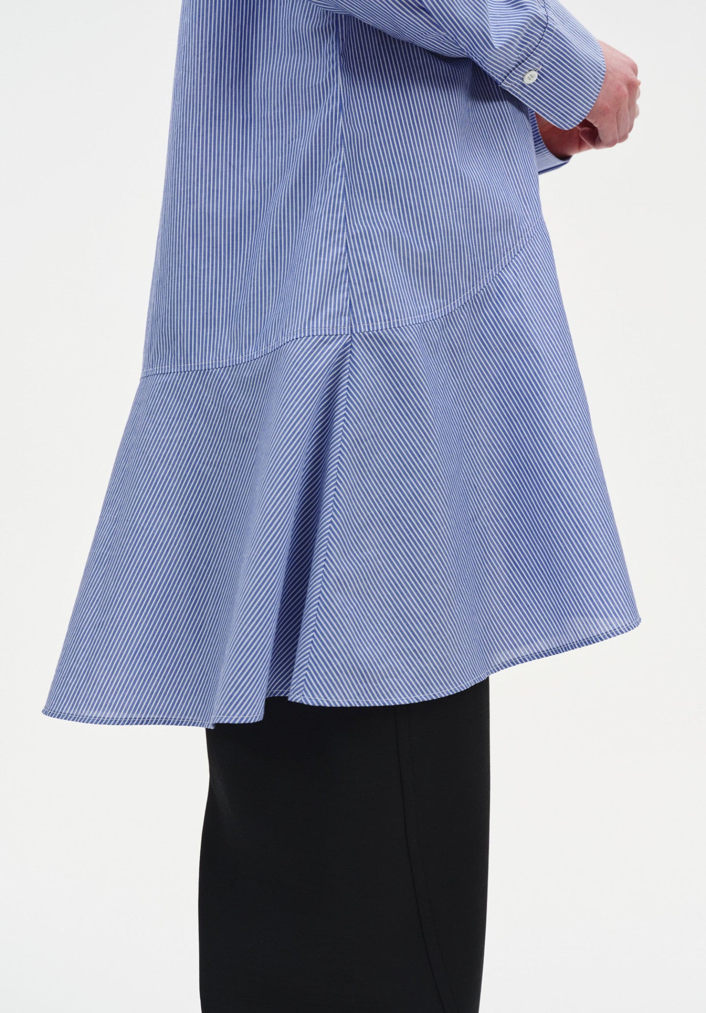 Close-up of a light blue long-sleeve tunic with fine vertical stripes, highlighting a curved seam and flared hem detail, worn over black wide-leg trousers.

