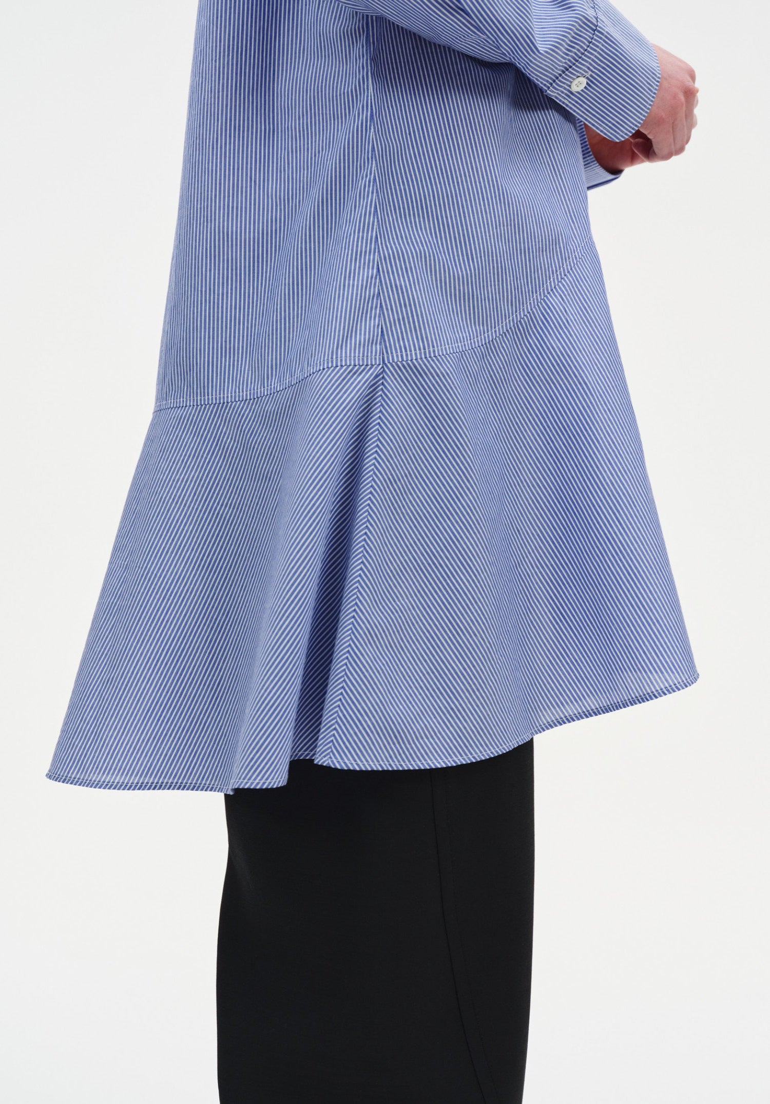 Close-up of a light blue long-sleeve tunic with fine vertical stripes, highlighting a curved seam and flared hem detail, worn over black wide-leg trousers.
