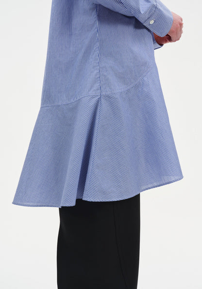 Close-up of a light blue long-sleeve tunic with fine vertical stripes, highlighting a curved seam and flared hem detail, worn over black wide-leg trousers.
