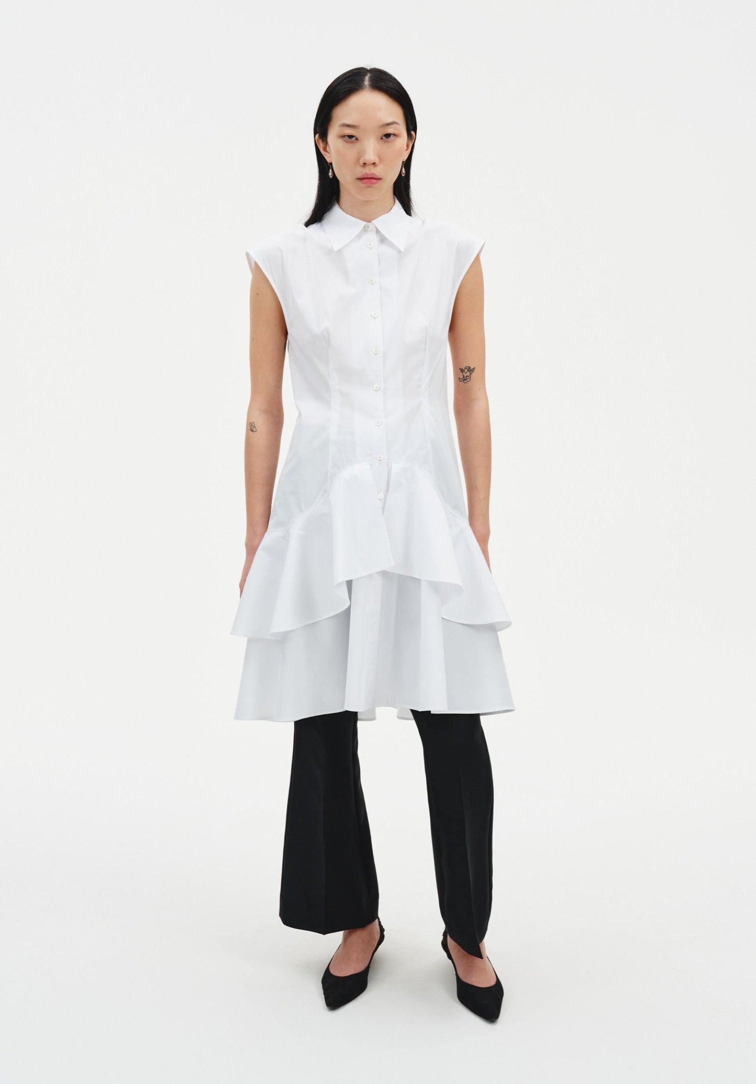 Front view of a model wearing a sleeveless white button-down tunic with a pointed collar and layered flared hem, styled over black slim trousers and paired with black pointed heels.
