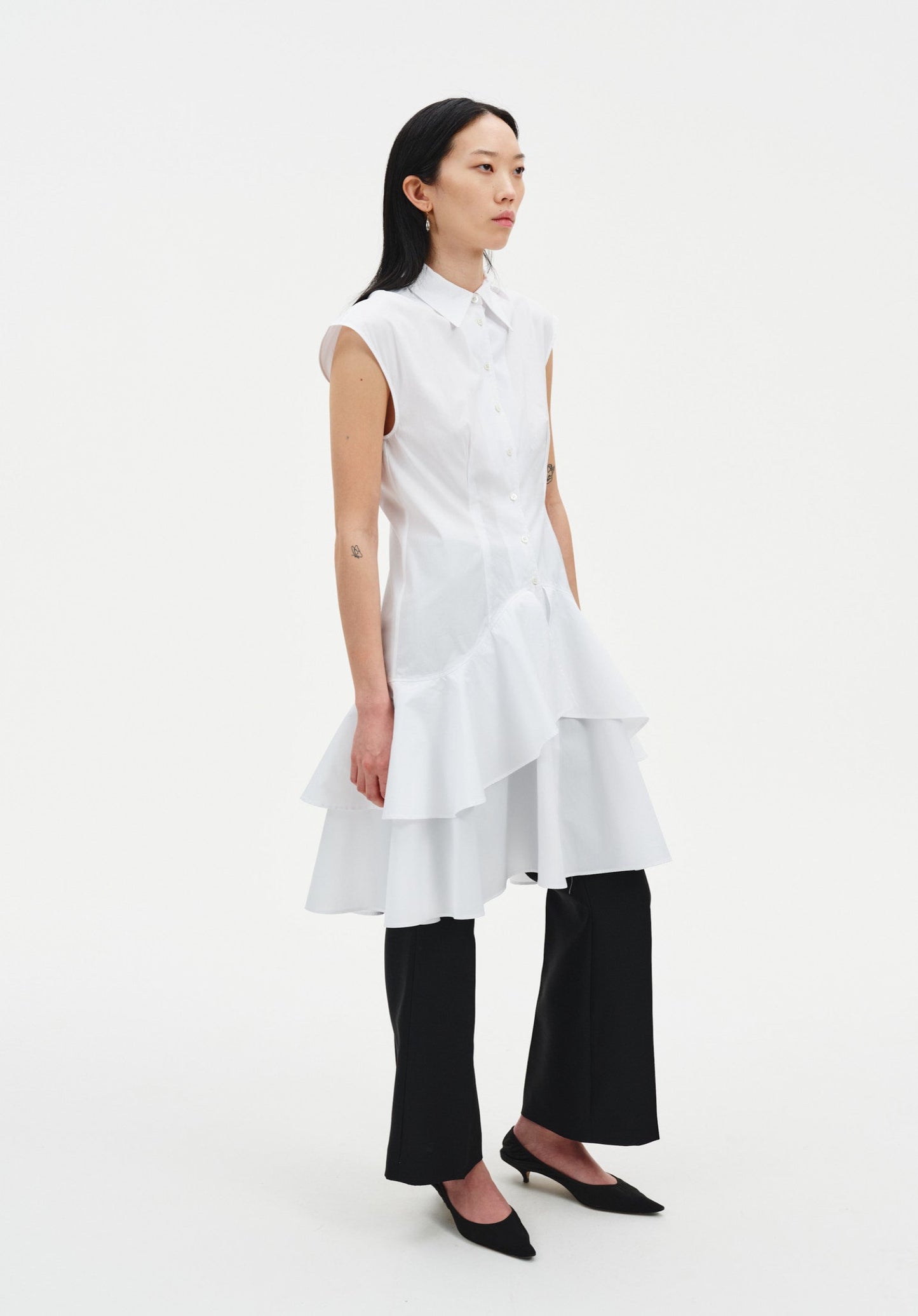 Side view of a model wearing a sleeveless white button-down tunic with a pointed collar and layered ruffled hem, styled over black cropped trousers and paired with black pointed heels.
