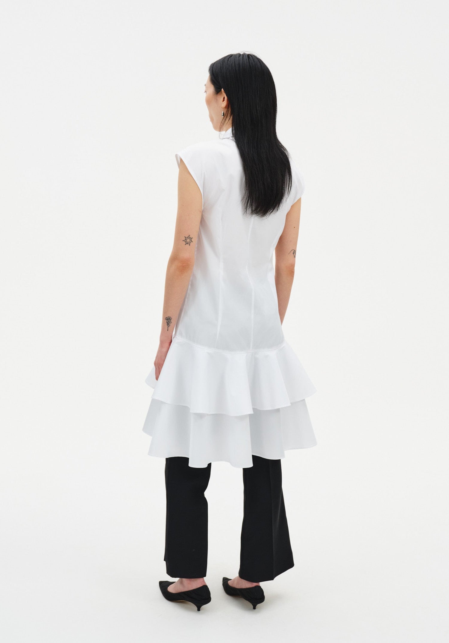 Back view of a model wearing a sleeveless white tunic with a structured fit and tiered ruffled hem, styled over black cropped trousers and paired with black pointed heels.
