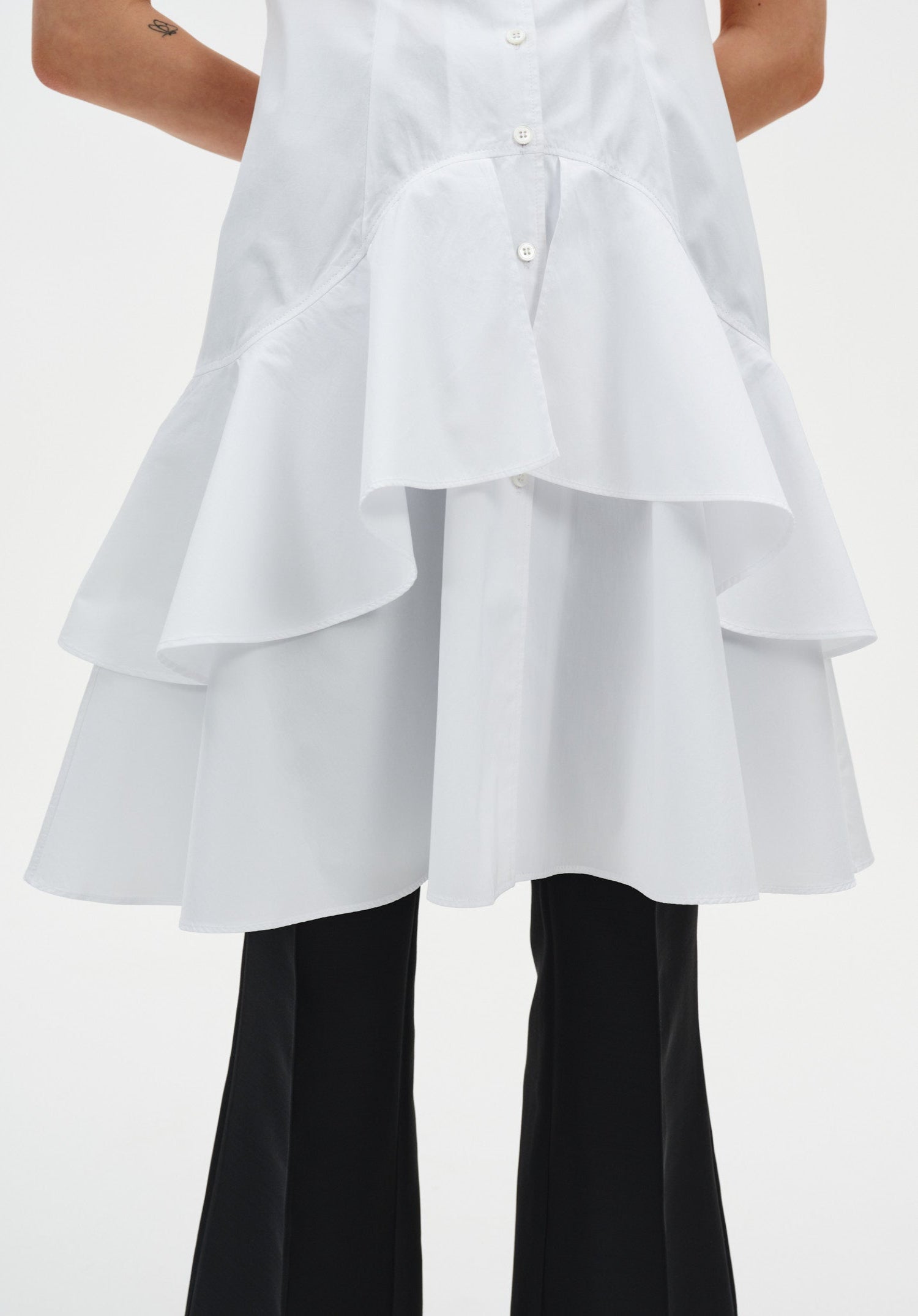 Close-up of a white sleeveless button-down tunic highlighting the layered, flared hem with soft ruffle tiers, worn over black trousers.
