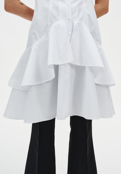 Close-up of a white sleeveless button-down tunic highlighting the layered, flared hem with soft ruffle tiers, worn over black trousers.
