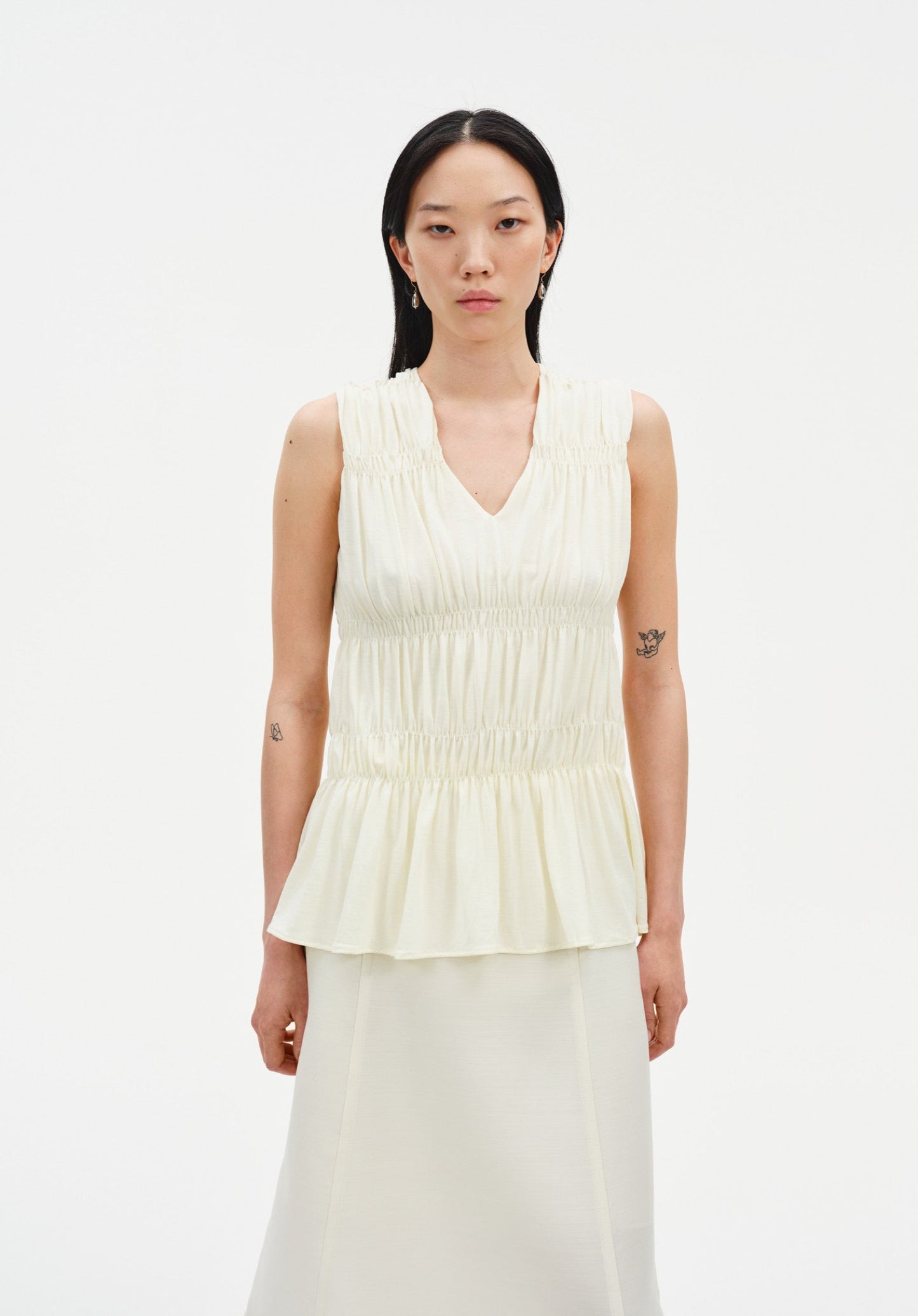 Front view of a sleeveless cream blouse with a V-neckline, gathered shirring, and peplum hem, paired with a matching skirt.
