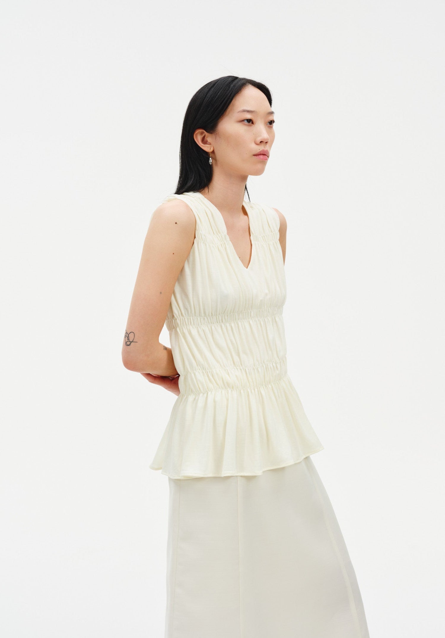 Side view of a model wearing a sleeveless cream blouse with a V-neckline, gathered shirring, and peplum hem, paired with a matching skirt.

