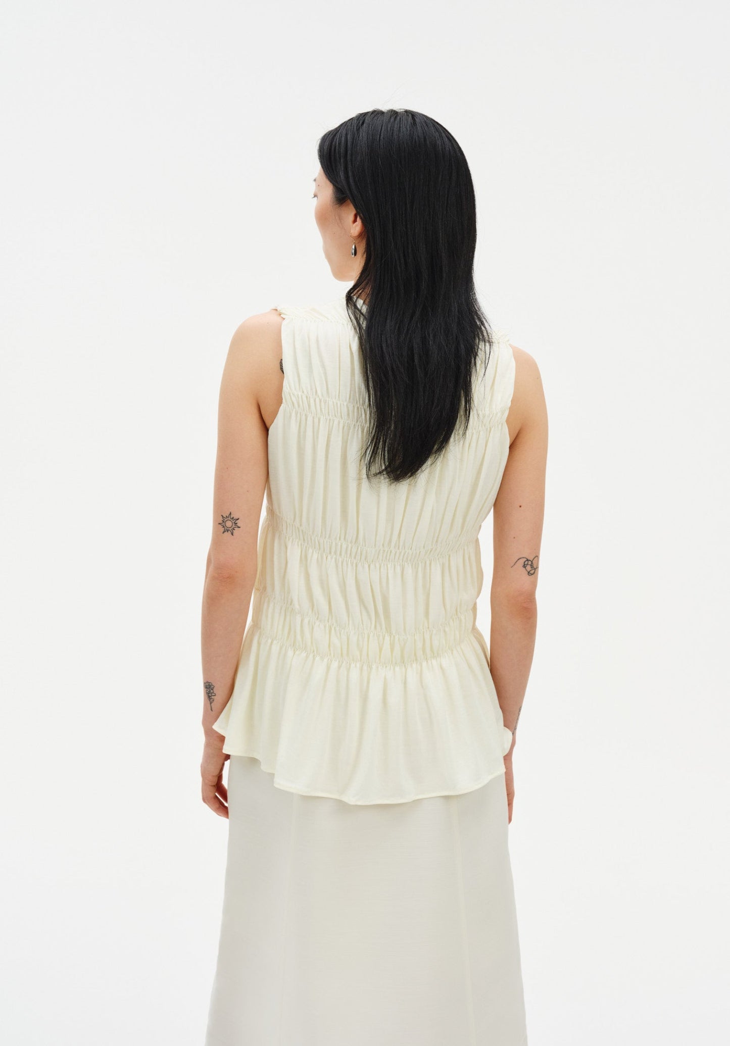 Back view of a light cream sleeveless top featuring soft, gathered tiers and a gently flared hem, highlighting its textured, flowy design against a plain background.
