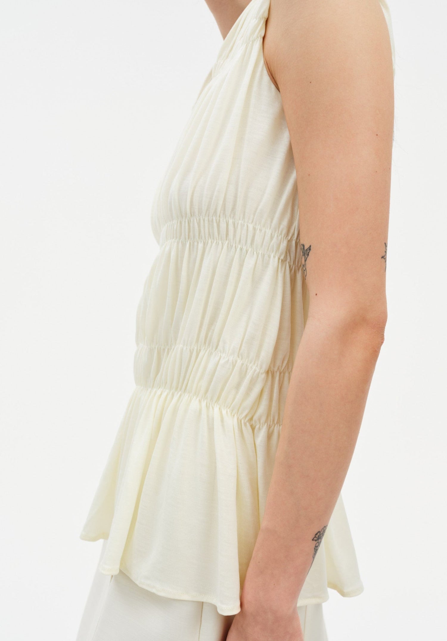 A light cream sleeveless top with gathered, tiered fabric; their arm is visible with small tattoos, and the background is plain and neutral.
